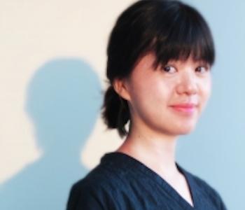 Profile image of Ying-Hsiu Chou in a dark blue top against a white wall with blue shadows. Her hair is dark and pulled back in a low bun with bangs in front. She is smiling close-lipped at the camera.