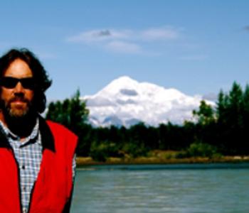 Richard Gray wears a red jacket and stands with a mountain in the background on a clear day.