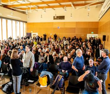 More than two hundred people sit or stand and talk with each other in a wood paneled room.