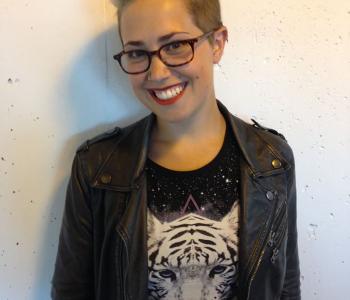 AJ smiles while wearing dark glasses a black shirt with a white tiger on it and a black jacket while standing in front of a white background.