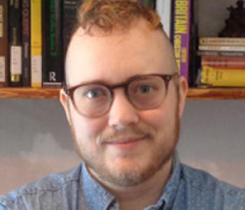 Adrian Kane wears glasses and a blue shirt with a bookcase in the background.