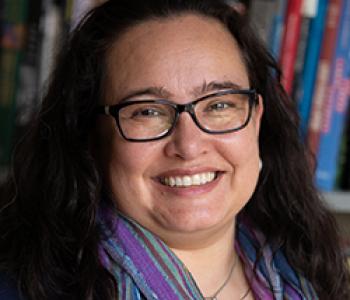 Portrait of Ariana Ochoa Camacho wearing glasses and standing in front of a bookcase.