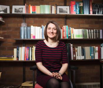 Profile of Beatrice in front of a book case. She has short brown hair and is wearing a sweater while smiling at the camera.