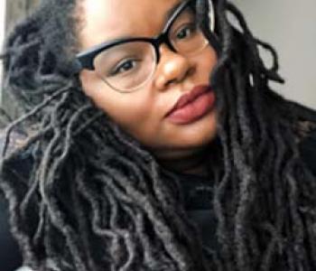 A close-up portrait of Bettina Judd wearing glasses and a dark shirt.