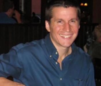 Nathaniel Bond wears a blue collared shirt while sitting in a chair and looking into the camera.