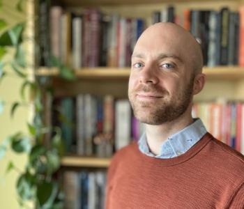 Caleb Knapp wearing blue oxford shirt and burnt orange sweater poses in front of bookshelf for portrait