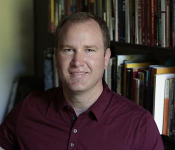 Christopher Teuton sits in front of a bookcase wearing a maroon shirt.