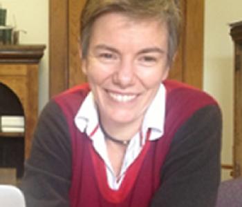 Cricket Keating sits behind a desk wearing a red and grey sweater.