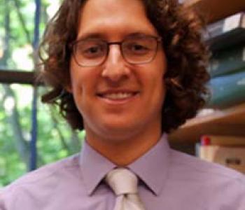 Portrait of Devin Naar wearing a purple shirt and tie and wearing glasses.