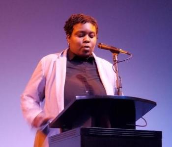 Picture of Rasheena at Hugo House on stage reading writing in front of microphone on at podium.