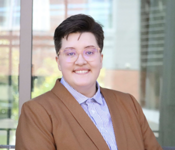 A white butch person with glasses smiling in a light brown blazer and blue and white checkered button-up shirt. 