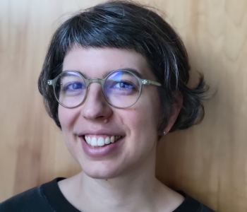 Portrait of Hannah Frydman standing in front of a brown wall wearing glasses.