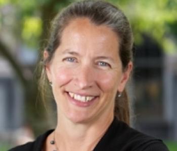 Sara Goering professional photo smiling at the camera, blurred trees in the background, wearing a blue blazer, necklace, and hair pulled back