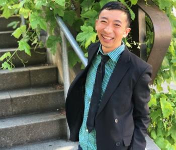 Douglas S. Ishii stands in front of stairs while wearing a blazer, teal collared shirt, and a black tie.