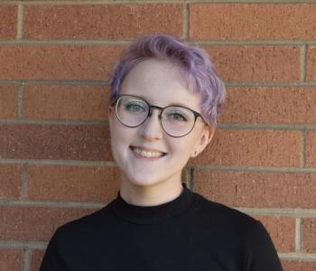 Head and shoulders of a smiling white person with short purple hair and glasses in a black turtleneck against a brick wall background