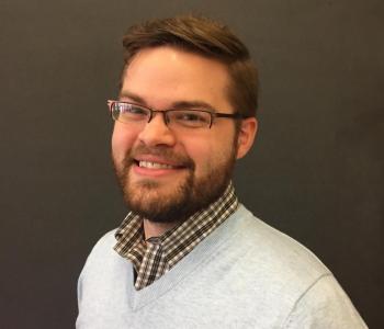 Justin Randolph stands in front of a dark wall while wearing glasses and a white sweater.