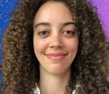 A profile picture of Kelly, a young woman with white skin and big brown curly hair, wearing a white collared shirt and smiling, with a blue to purple gradient background with glitter sparkles.