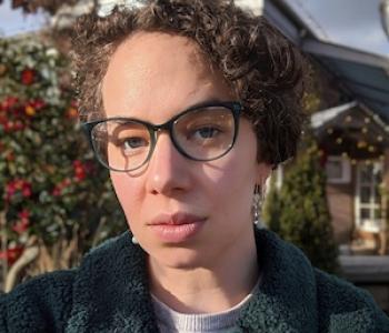 profile of Kaitlyn Boulding in glasses, close-up to the camera, wearing a jacket with a sky, trees, and buildings blurred in the background