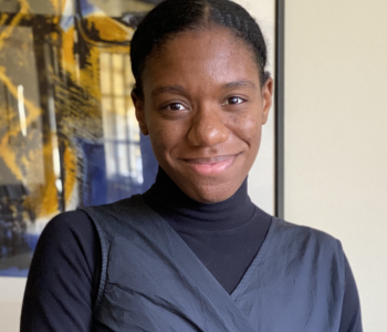 profile of Kalia smiling at the camera, hair back in a bun and in a black turtle neck top with overlapping black dress