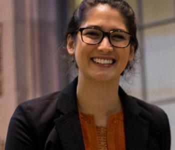 Profile of Katia smiling at the camera in a dark blazer and orange shirt