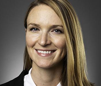 Kristin Dew stands in front of a dark wall wearing a white shirt and dark jacket.