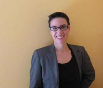 Picture of Sarah Levin-Richardson standing in front of a beige wall wearing glasses, a blazer, and a black shirt.