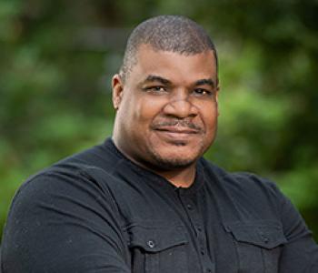 A portrait of Marcus Johnson standing in front of tress wearing a black shirt.