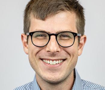 Matthew Bellinger stands in front of a white wall wearing a patterned shirt and glasses.