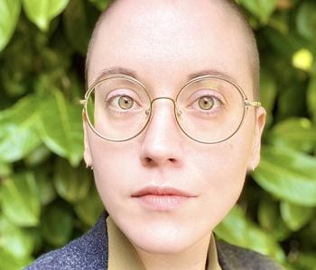 Maxine Savage stands in front of a large bush while wearing glasses and a grey jacket.