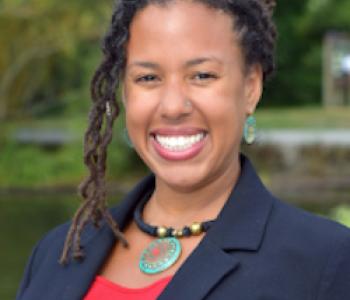 profile of Maya Smith smiling at the camera, hair pulled back with some curls along the left side, wearing a red top under a blue blazer with a necklace