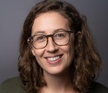 Headshot of Melanie Walsh standing in front of a grey wall wearing glasses.