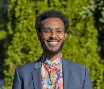 Nathanael in front of a backdrop of green foliage, smiling while wearing a blue blazer and floral shirt.