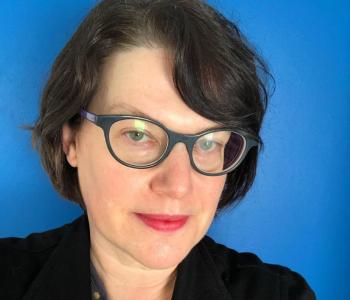 Portrait of Leigh Mercer wearing glasses and a dark jacket standing in front of a blue wall.