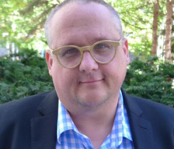 Portrait of Michael Blake wearing a suit and sitting outside.