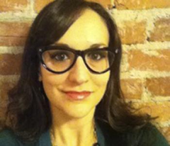 Pamela Pietrucci stands in front of a brick wall wearing glasses an a green shirt.