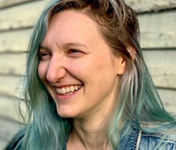 Jacqui Pratt has blue hair and looks to the left and laughs while standing in front of a shiplapped exterior wall.