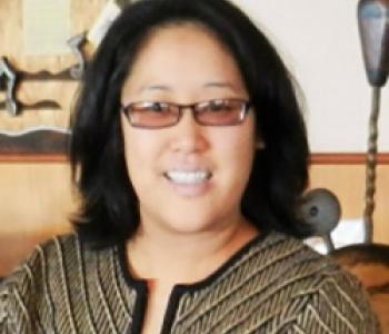 Portrait of Regina Yung Lee wearing glasses and sitting in front of a light wall.