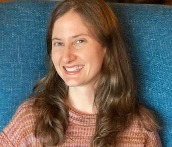 Adair Rounthwaite sits on a blue chair wearing a pink sweater. 