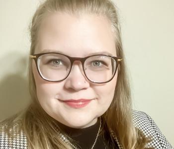 Samantha Thompson stands in front of a beige wall while wearing glasses and a jacket.