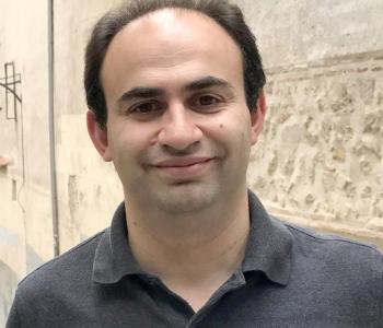 Sasha Senderovich stands in front of a wood panel while wearing a dark collared shirt.