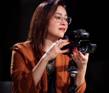 Sonia De La Cruz looks to the right while standing behind a camera on a tripod.