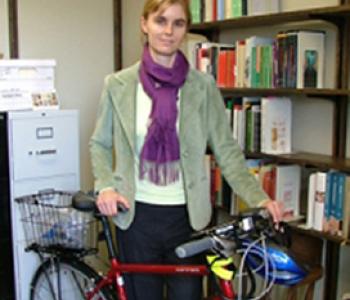 Susan Gaylard stands behind a red bicycle and in front of a bookcase wearing a light-green jacket and purple sweater.