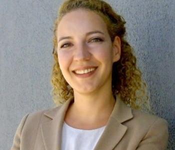 Ayda smiling at the camera in a beige blazer and white shirt, curly hair pulled halfway back and a gray background