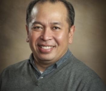 professional photo of Rick Bonus smiling at the camera in a blue shirt and a brown background