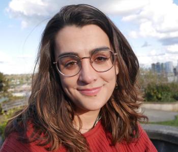 Close-up photo of a smiling woman with brown hair wearing round glasses and a red sweater. Part of the Seattle skyline is in view behind her.