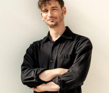 James Pierce stands in front of a white wall with his arms crossed while wearing a black collared shirt and black trousers.