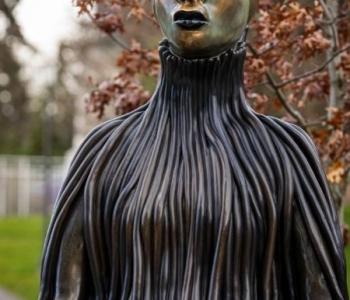 bronze statute of an African Women with an obelisk over her eyes and forehead, a garmetn in ribbons running down her seated body, hands on her knees