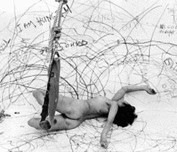 Photograph of Carolee Schneemann naked drawing on a room-sized paper and attached to a swing.