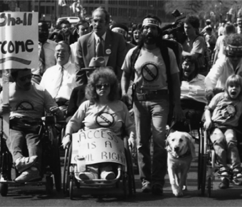 A black and white photo of a group of people walking and in wheelchairs protesting with signs.