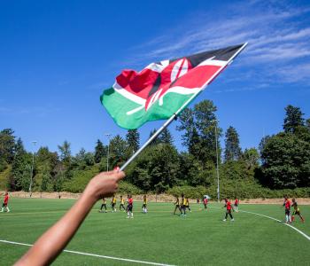 Hand holding a Kenyan flag with a scoccer field in the background